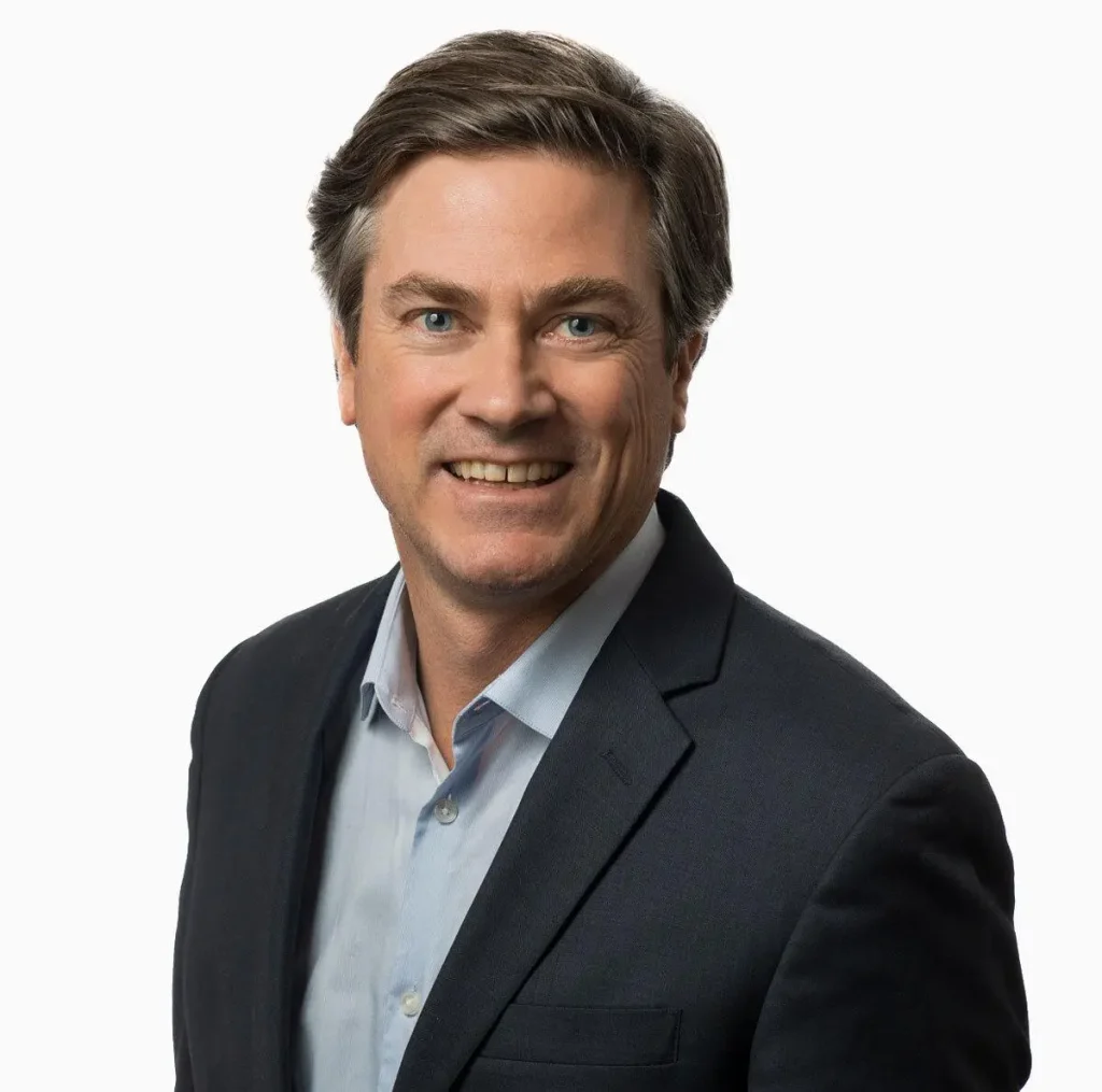 John Wakefield smiling in a professional portrait, wearing a dark blazer over a light blue shirt against a plain white background.