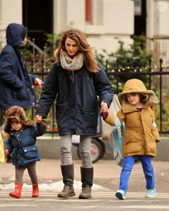 Keri Russell walks across a city street holding hands with her children River and Willa, who are dressed in winter clothing.