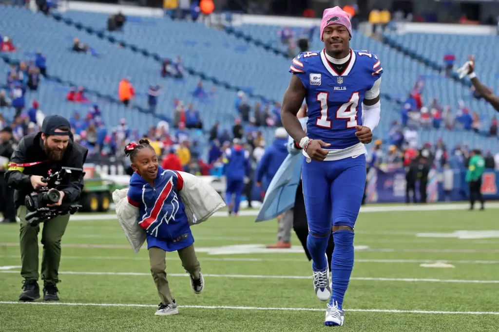Stefon Diggs running on the football field alongside his daughter, Nova, who is wearing a Buffalo Bills outfit.