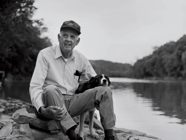 Wendell Berry sitting on rocks beside a river with his dog, wearing a cap and light-colored shirt and trousers.