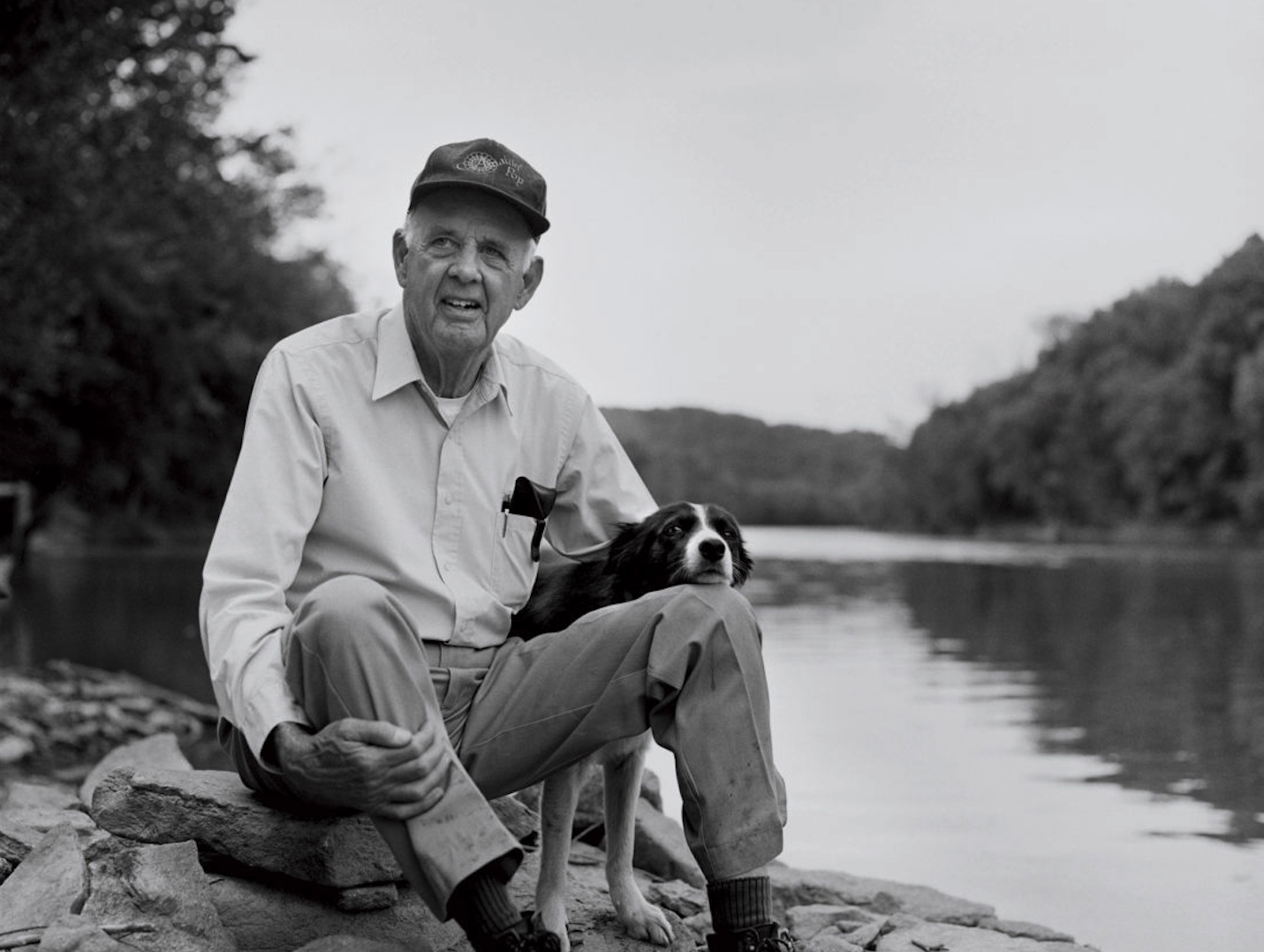 Wendell Berry sitting on rocks beside a river with his dog, wearing a cap and light-colored shirt and trousers.