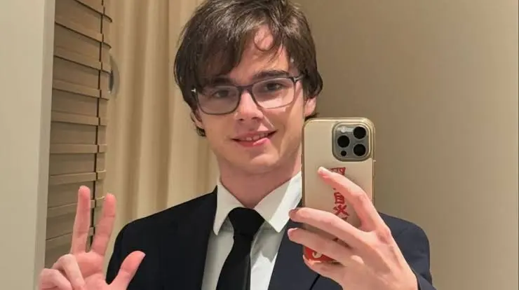 Valentino Martin wearing a black suit, white shirt, and black tie while taking a mirror selfie with a smartphone.
