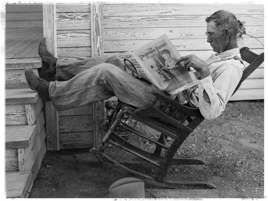 Wendell Berry sitting in a rocking chair outside a rustic wooden home, reading a copy of the Progressive Farmer magazine.