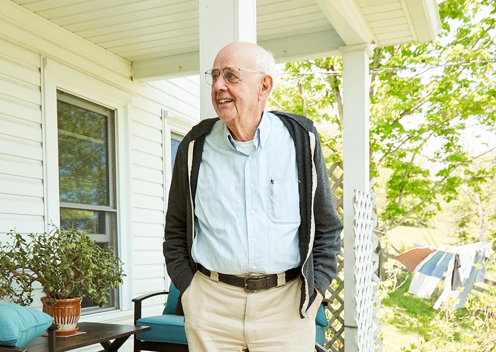 Wendell Berry standing on a porch outside his home, wearing glasses, a light blue shirt, dark cardigan, and beige pants.