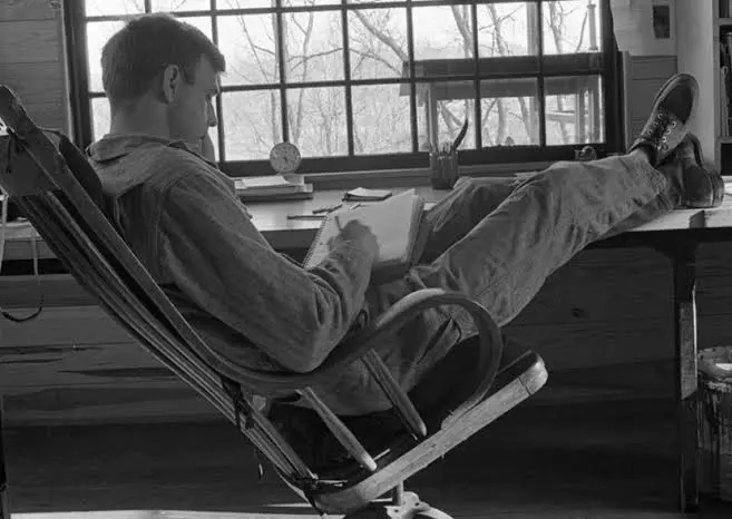 Wendell Berry writing in a notebook while reclining in a chair beside a large window.