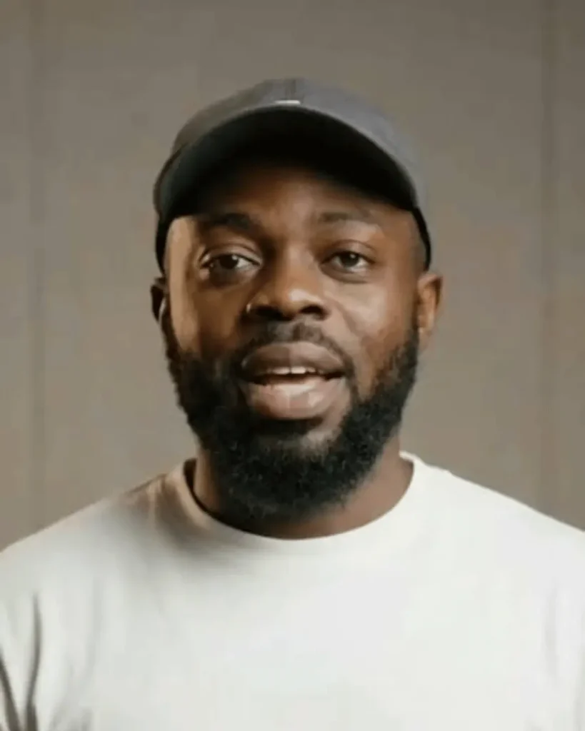 Dave Chappelle speaking in a video interview while wearing a black cap and white shirt