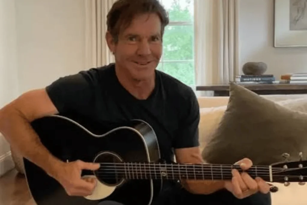 Dennis Quaid playing an acoustic guitar while sitting indoors