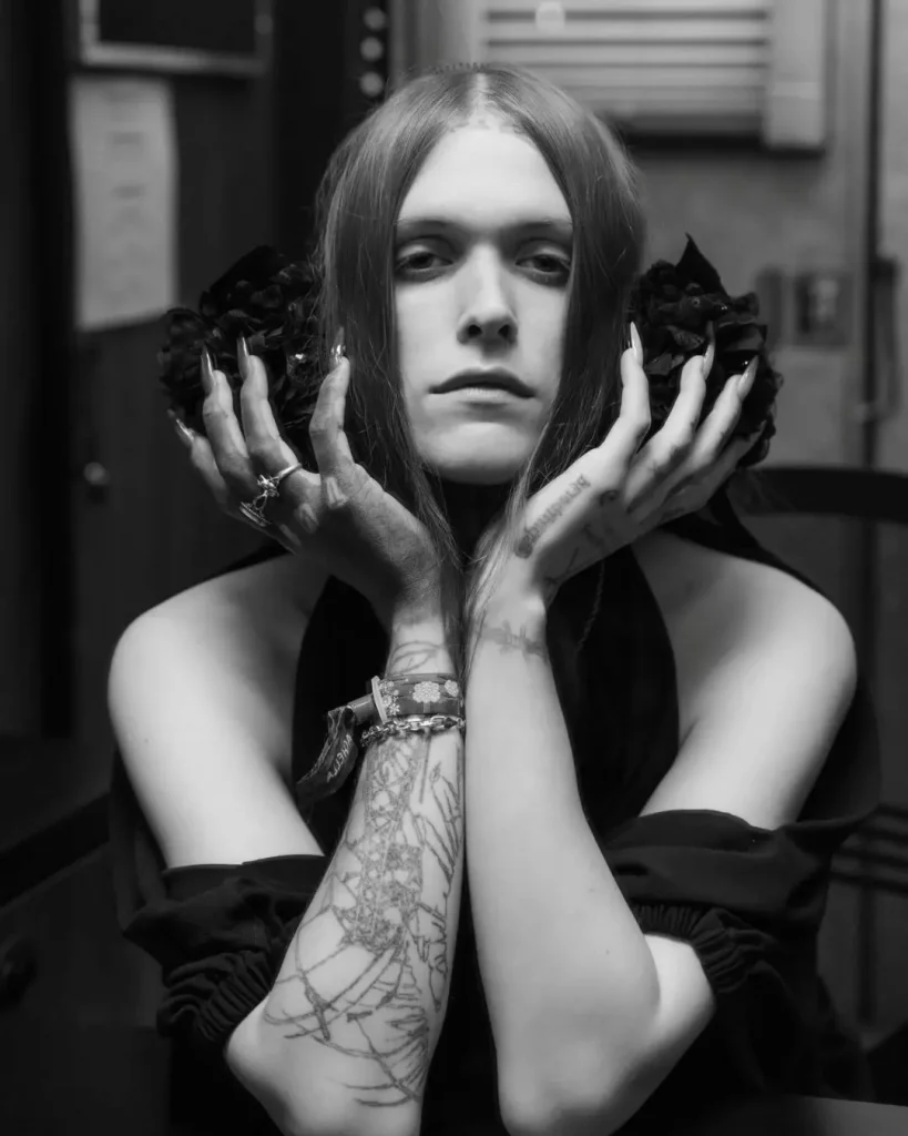 Black and white close-up portrait of Ethel Cain holding her face with tattooed hands, featuring dark hair and floral arrangements.