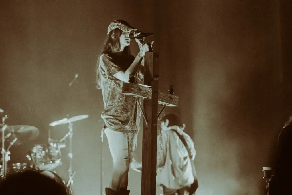 Ethel Cain performing on stage wearing a cap and t-shirt, singing into a microphone near a wooden cross structure.