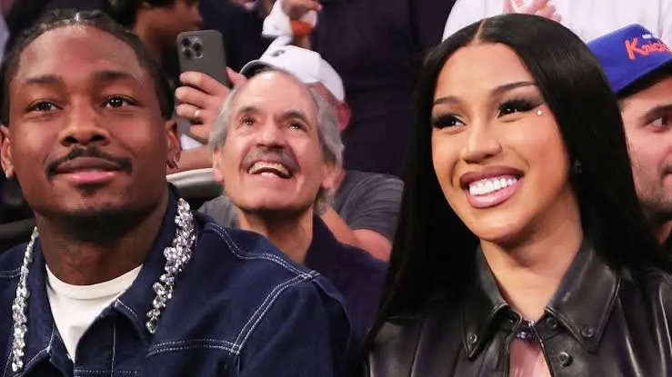 Cardi B and Stefon Diggs smiling and posing together at a basketball game, seated courtside with a cheering crowd in the background.