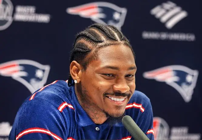 Stefon Diggs with braided hair smiles during a press conference while speaking into a microphone, wearing a navy blue and red sports polo shirt.