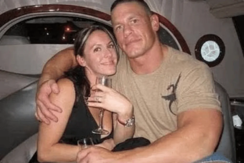 John Cena with his arm around Elizabeth Huberdeau holding a glass inside a cabin or boat with round windows.
