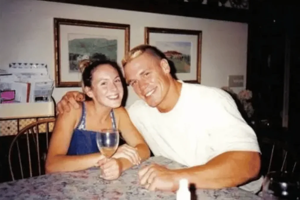 John Cena with his arm around his first wife Elizabeth Huberdeau holding a wine glass while sitting at a dining table.
