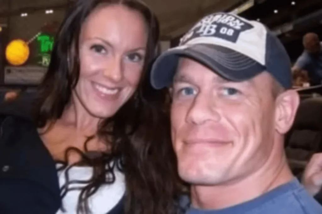 John Cena wearing a baseball cap alongside Elizabeth Huberdeau smiling together at a crowded sporting venue.