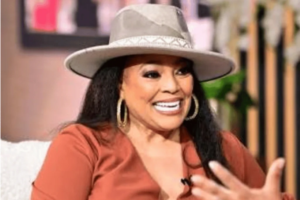 Kim Fields wearing a styled fedora hat and rust-colored top while speaking during an interview.