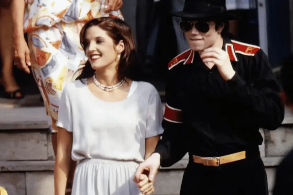 Lisa Marie Presley wearing a white outfit and pearl necklace holding hands with Michael Jackson, who wears a black outfit with red accents and aviator sunglasses.