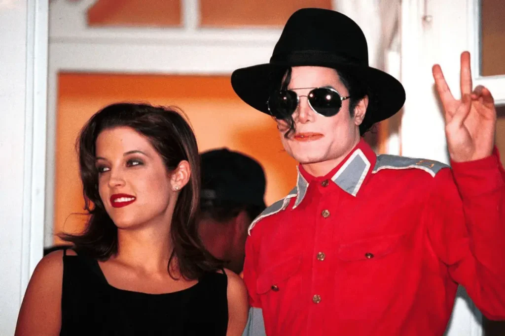 Lisa Marie Presley in a black top standing beside Michael Jackson in a bright red collared shirt, hat, and sunglasses flashing a peace sign.