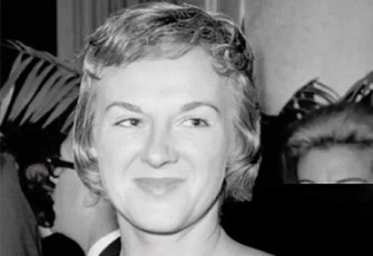 Black-and-white portrait of Margie Willett smiling softly while looking slightly to the side, with people and indoor foliage visible in the background.